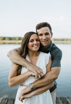 a man and woman hugging each other on a dock by the water with their arms around each other