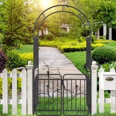 an iron gate in front of a white picket fence with green grass and bushes surrounding it