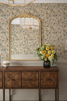 a vase with flowers on top of a wooden table in front of a wallpapered mirror