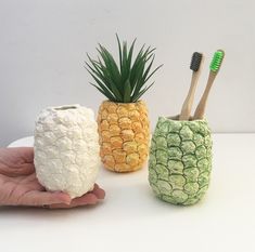 three pineapple shaped toothbrush holders in different shapes and sizes, with one holding a bamboo toothbrush