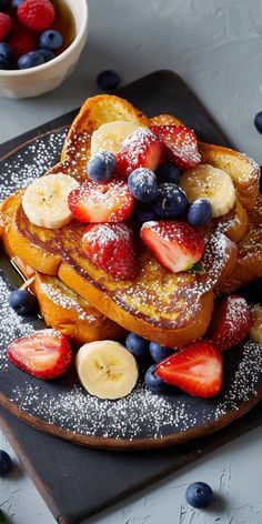 french toast topped with fresh fruit and powdered sugar on a black plate next to a bowl of berries