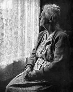 an old woman sitting in front of a window looking out at the outside with lace curtains