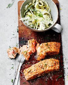 two pieces of meat on a cutting board next to a bowl of pasta and a fork