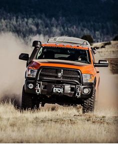 an orange truck driving down a dirt road in the middle of a field with dust behind it