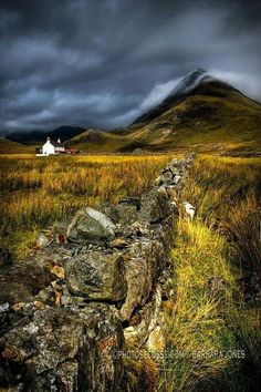 a stone wall in the middle of a grassy field with mountains in the background under a cloudy sky