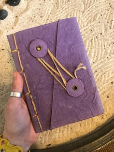 a hand holding a purple piece of paper with string and buttons attached to it, on top of a table