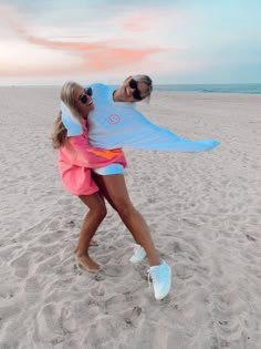 two people standing on top of a sandy beach near the ocean with one person wearing sunglasses