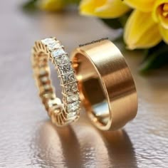 two gold wedding rings sitting next to each other on top of a table with flowers in the background