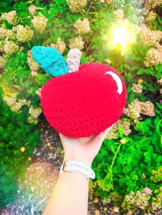 a hand holding an apple shaped object in front of some bushes and flowers with the sun shining on it