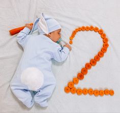 a baby laying on top of a white bed next to orange beads and carrots