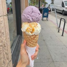 a hand holding an ice cream cone with purple and white toppings on it in front of a sidewalk