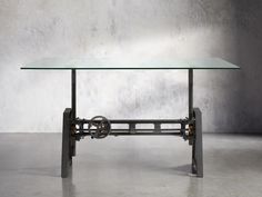 a glass table with metal frame and wheels on the bottom, in front of a concrete wall