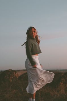 a woman standing in the middle of a field wearing a skirt and hoodie with her hair blowing in the wind