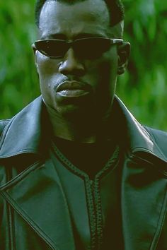 a man wearing sunglasses and a leather jacket looks at the camera while standing in front of trees