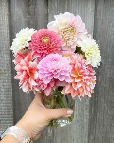 a person holding a glass vase filled with pink, white and yellow flowers on top of a wooden fence