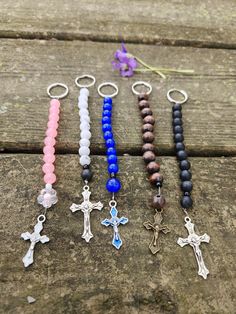 six different colored beads and cross charms on a wooden table next to a purple flower