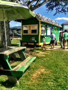 people are standing around in front of small green buildings with picnic tables and umbrellas