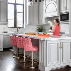 a woman standing in a kitchen next to a counter with pink chairs and white cabinets