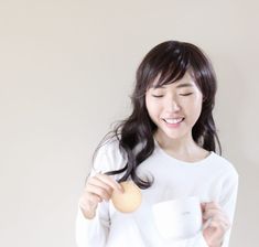 a woman holding a cup and a doughnut in her right hand while standing next to a white wall