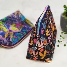 two zippered pouches sitting next to each other on a white surface with plants in the background