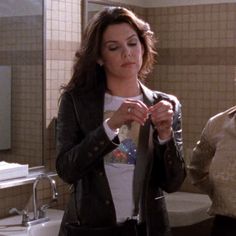 two women standing in a bathroom and one is tying her jacket while the other looks on