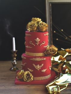 a three tiered red cake with gold roses on top and candles in the background