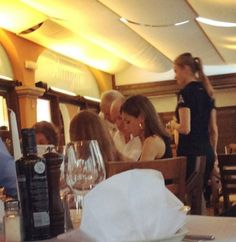 several people sitting at a restaurant table with wine glasses on the table and one person standing in front of them