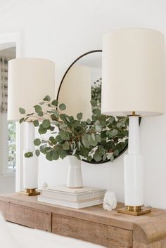 two white lamps sitting on top of a wooden table next to a mirror and lamp shade