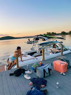 two people sitting on the dock next to a boat with luggage and water sports equipment