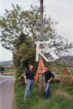 two men standing next to a wooden sign on the side of a road with trees in the background