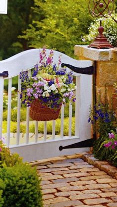 a white gate with flowers hanging from it's sides and a brick walkway leading to the front door