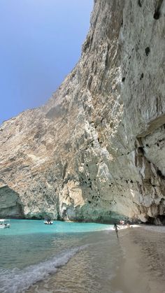 people are walking on the beach next to some cliffs and boats in the blue water