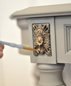 a person is painting the side of a white cabinet with gold decorations on it and holding a paintbrush in their hand