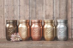 four gold, silver and copper mason jars are lined up on a wooden surface with a flower in the middle