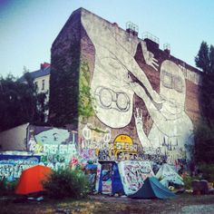 graffiti on the side of a building with tents set up next to it and trees in the background