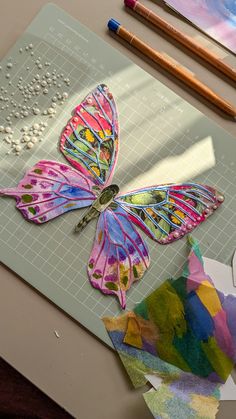 a paper butterfly sitting on top of a cutting board next to some colored pencils