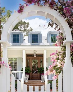 the entrance to a white house with pink flowers on it's front gate and an arched wooden door