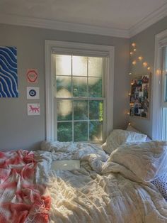 an unmade bed with blankets and pillows in front of a window that has string lights on it