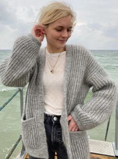 a woman is standing on a dock by the ocean wearing a gray cardigan sweater and jeans