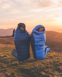 two people are sitting on the grass with their feet up and one is wearing a sleeping bag