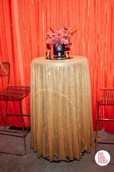 a gold table cloth with flowers on top and two chairs around it in front of a red curtain