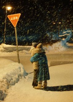 two people standing next to each other in the snow near a street sign and road