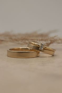 two gold wedding rings with a single diamond on each one, sitting next to some dried grass