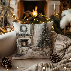 a christmas scene pillow sitting on a couch in front of a fire place with pine cones