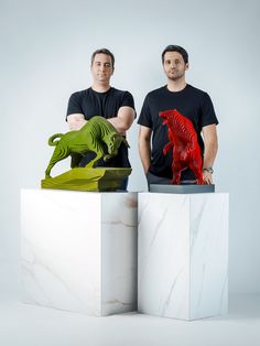 two men standing next to each other with sculptures in front of them on pedestals