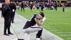 a football player kneeling down on the sidelines with his head in his hands and other players standing behind him