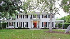 a large white house with black shutters on the front and red door, surrounded by trees