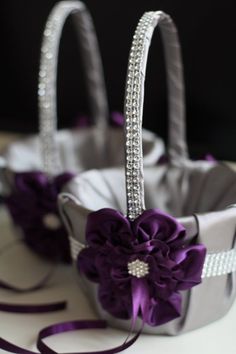 two silver baskets with purple flowers in them on a white tablecloth and ribbon tied around the handles