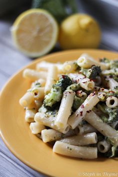 a yellow plate topped with pasta and broccoli covered in sauce next to lemon wedges