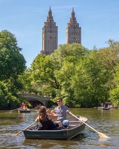 two people in a row boat on the river with tall buildings in the background,